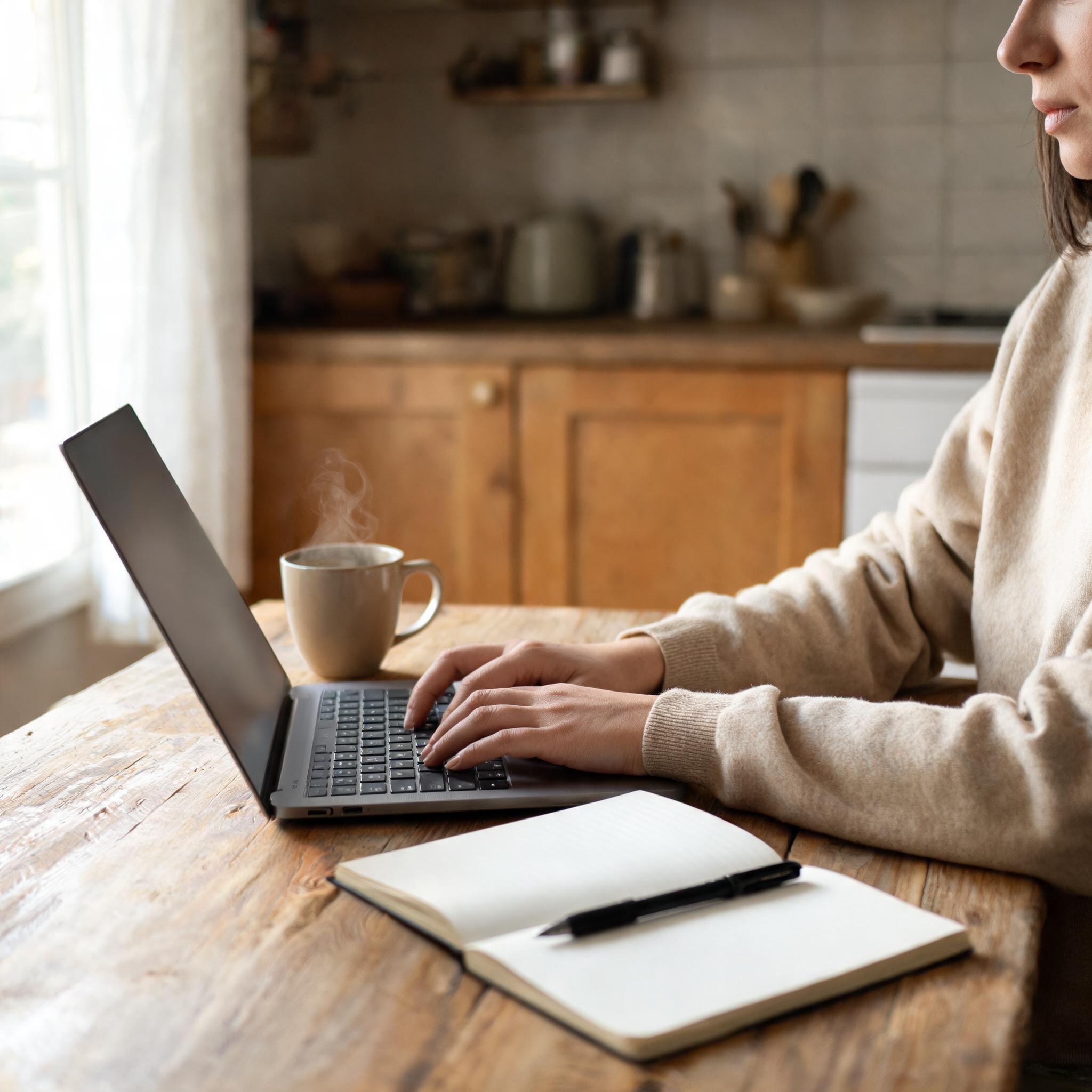 Person typing on a laptop at a wooden table with coffee and an open notebook nearby